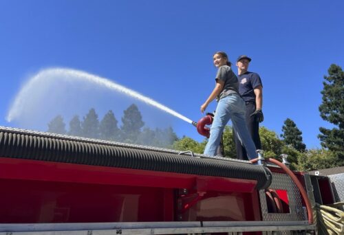 Fire And Emergency Services Teacher With Student Sparying Water Off Firetruck