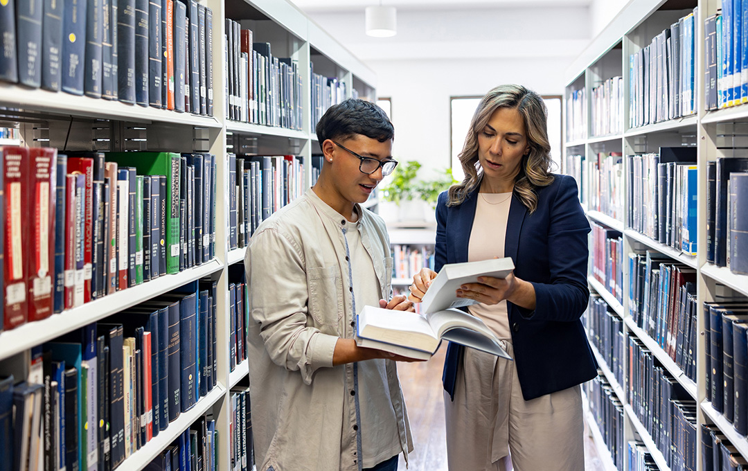 A student and an instructor review books together in a library aisle lined with shelves of academic resources.