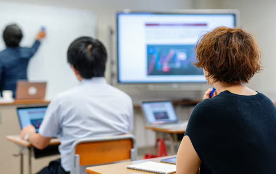 Classroom setting with adult learners using laptops while an instructor teaches at the front of the room beside a large presentation screen.