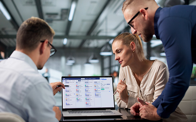 Three Colleagues Review A Project Dashboard Together On A Laptop During A Collaborative Planning Session.