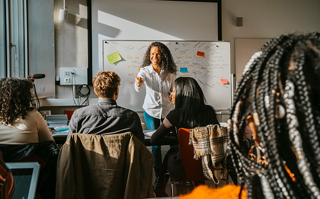 An Instructor Leads A Classroom Discussion While Standing In Front Of A Whiteboard As Students Listen And Participate.