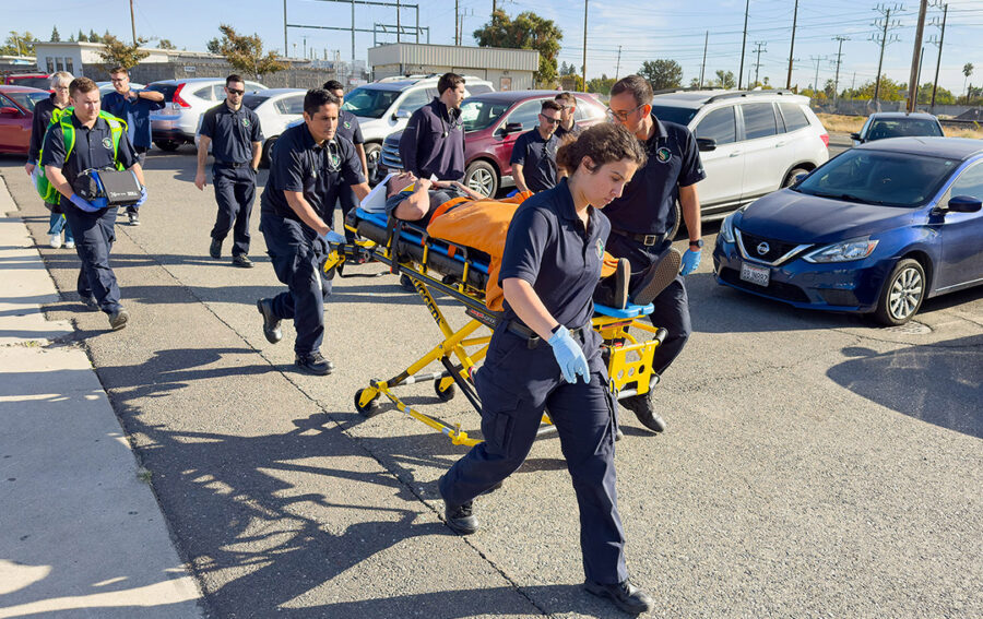 Paramedic students in uniform conduct a hands-on emergency response training exercise, transporting a simulated patient on a stretcher through a parking lot.