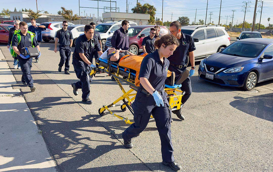 Paramedic students in uniform conduct a hands-on emergency response training exercise, transporting a simulated patient on a stretcher through a parking lot.