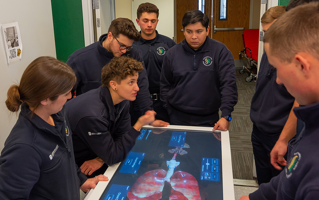 Paramedic students gather around an interactive anatomy table, studying a digital visualization of the human respiratory system during hands-on training.