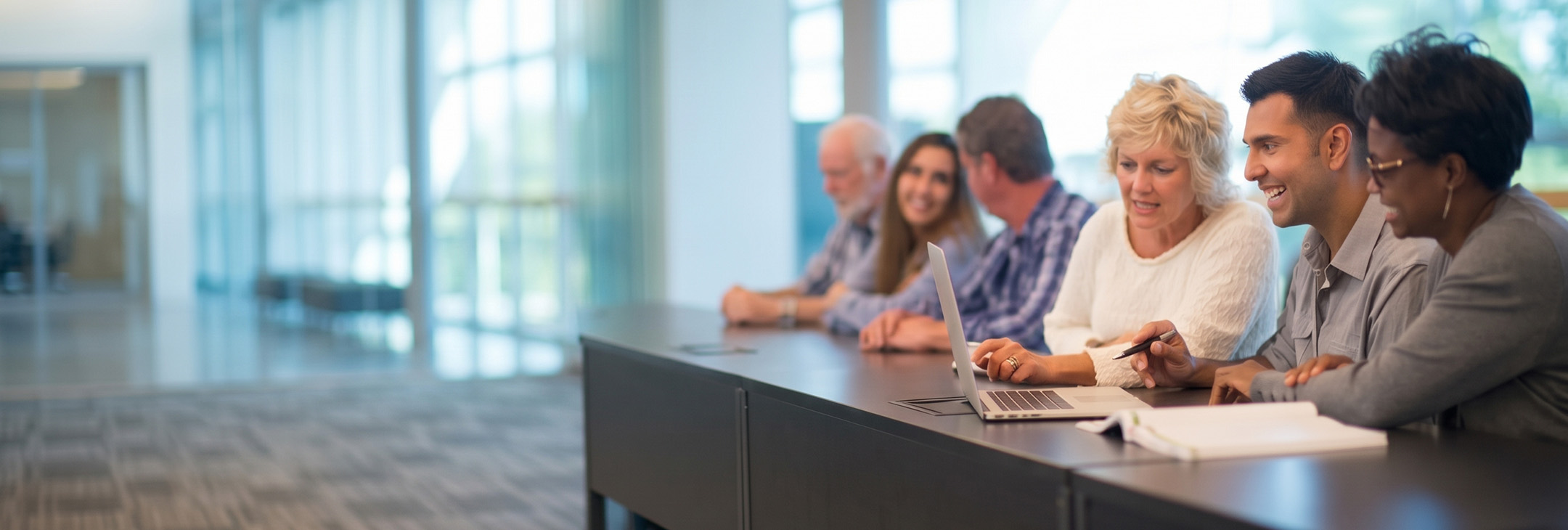 Adult students collaborate around a laptop in a modern classroom.