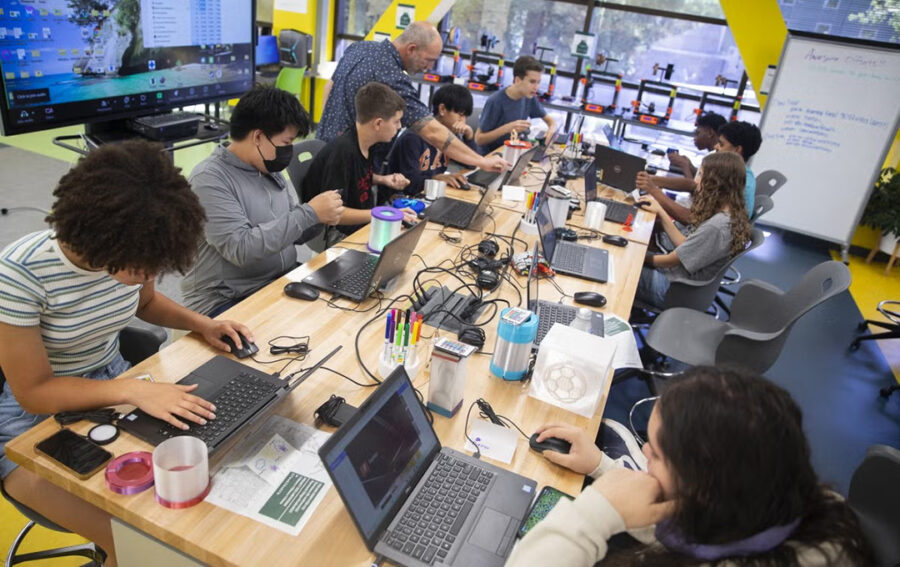 Students work on laptops and hands-on projects with an instructor in a classroom equipped with 3D printers.