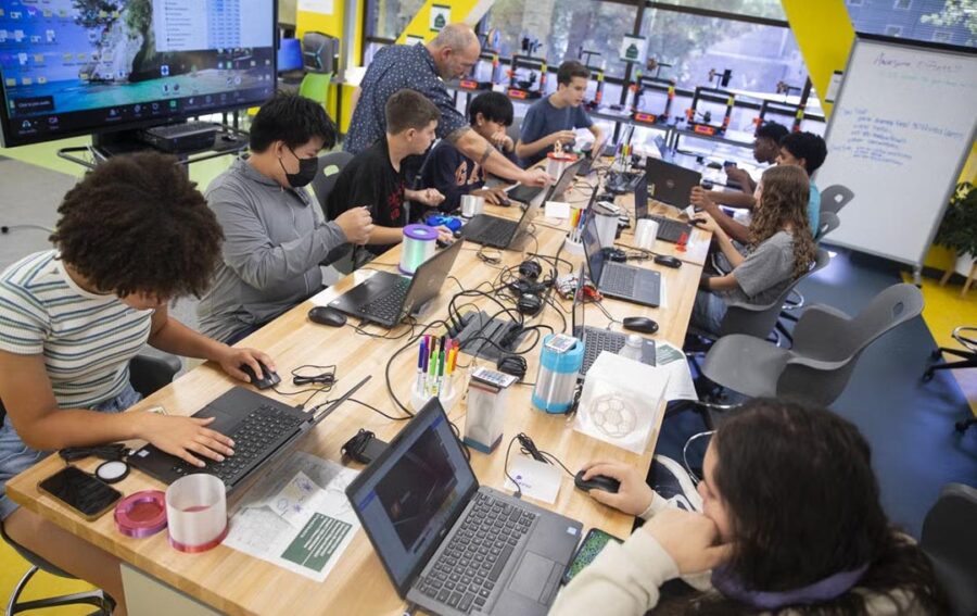 Students work on laptops and hands-on projects with an instructor in a classroom equipped with 3D printers.