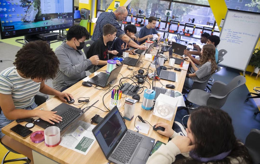 Students work on laptops and hands-on projects with an instructor in a classroom equipped with 3D printers.